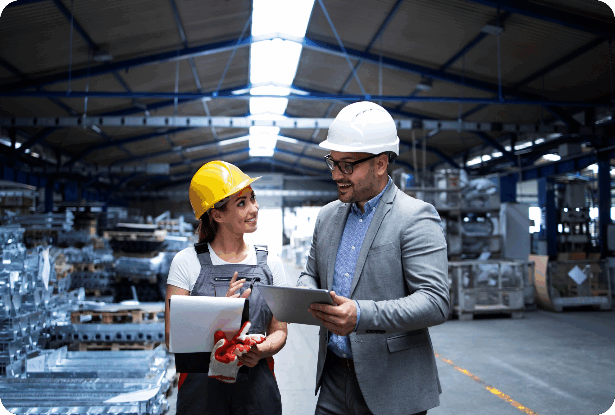 Man in a suit and hard hat discussing production plans with co-worker wearing safety gear and holding documents inside an industrial warehouse.