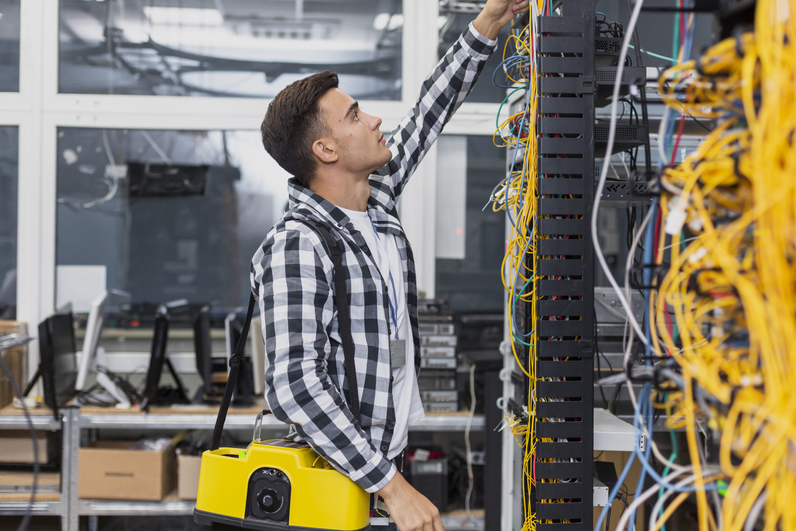 Technician working in a server room, organizing network cables connected to a rack while carrying testing equipment.